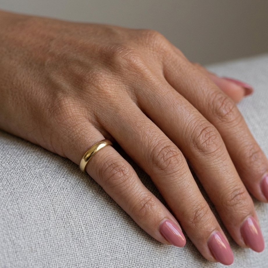 Hand wearing a gold ring with pink nail polish on a neutral background