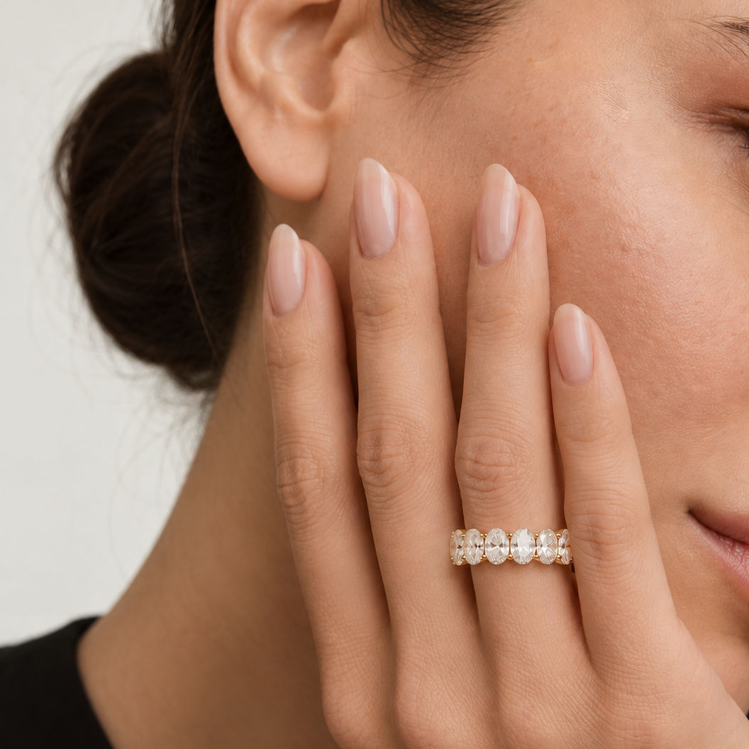 A close-up lifestyle shot of a woman's hand resting against her cheek, wearing a delicate gold ring adorned with small, clear teardrop-cut gemstones.