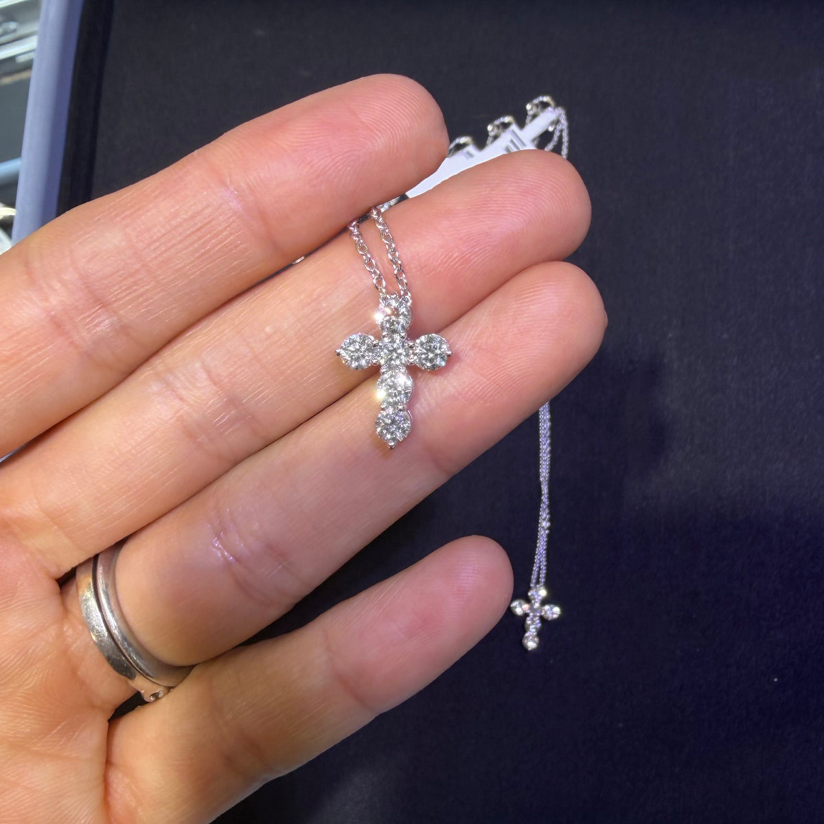 A silver cross pendant necklace with clear stones, held in a person's hand over a dark jewelry box.