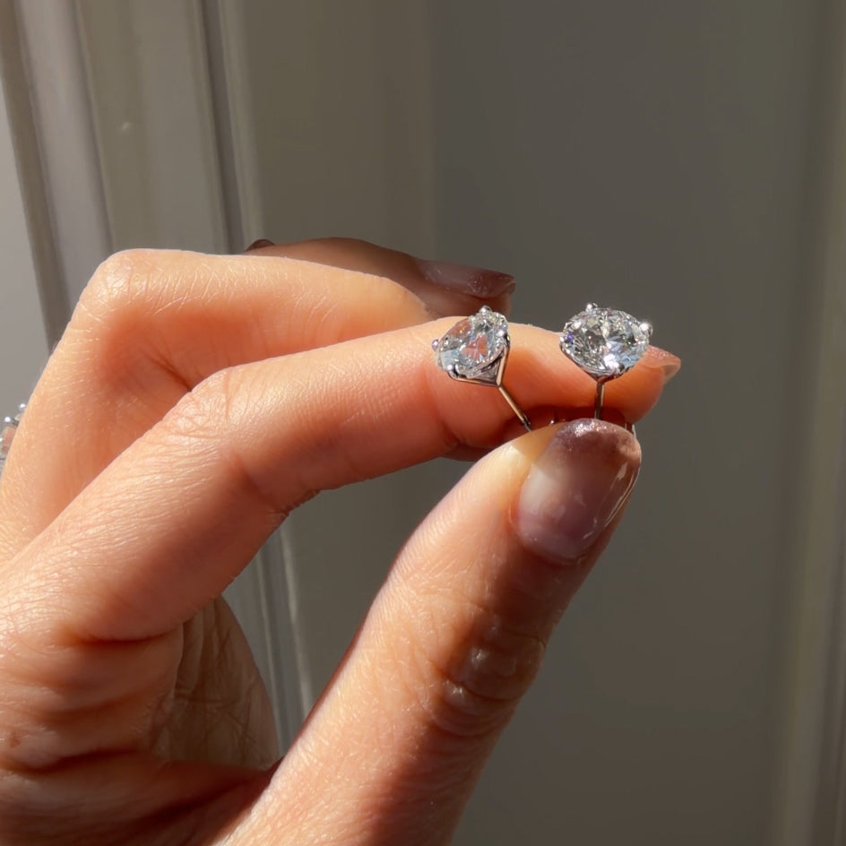 Hand holding two diamond stud earrings against a neutral background