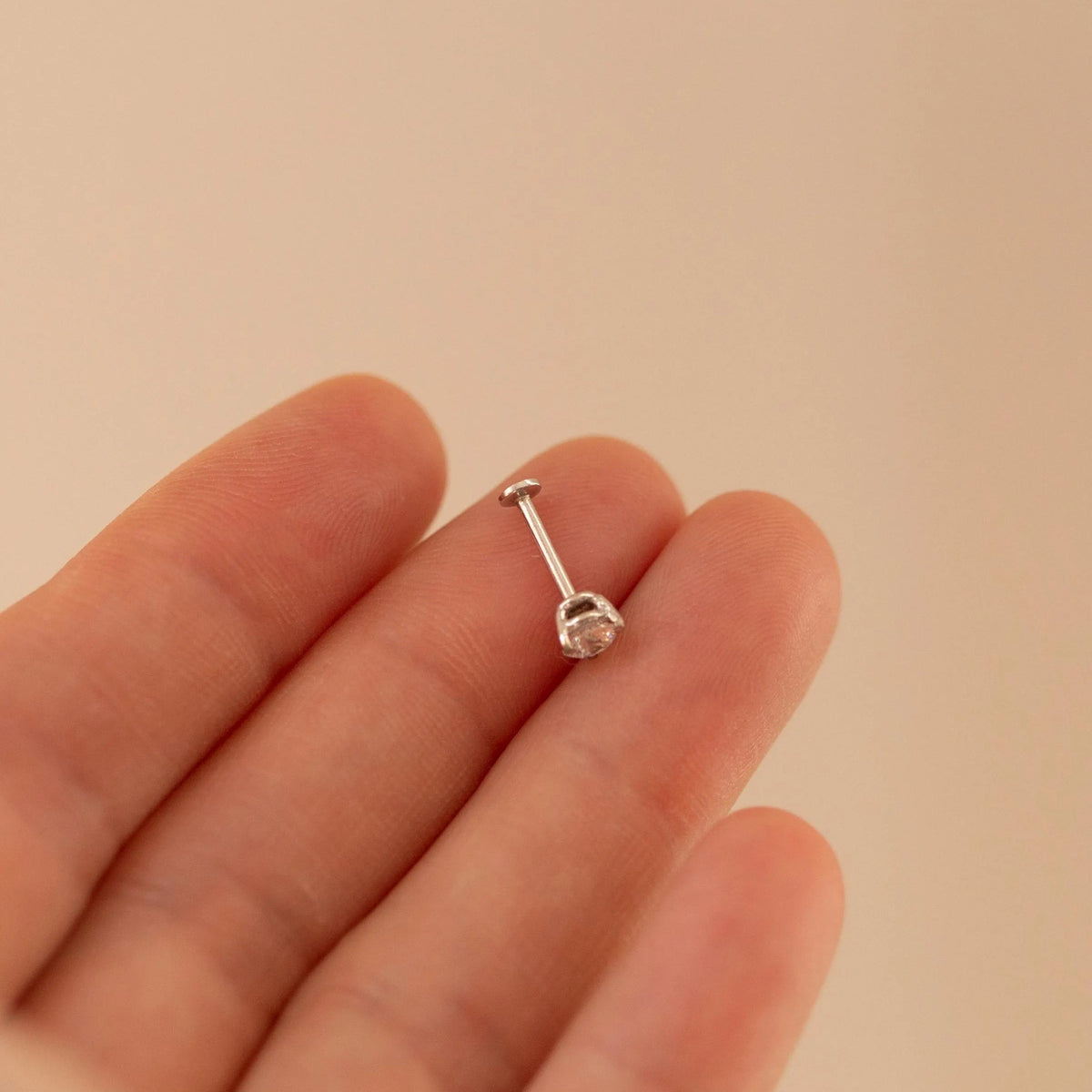 A silver-toned labret piercing stud with a clear gemstone, shown resting on a person's fingers against a soft beige background.