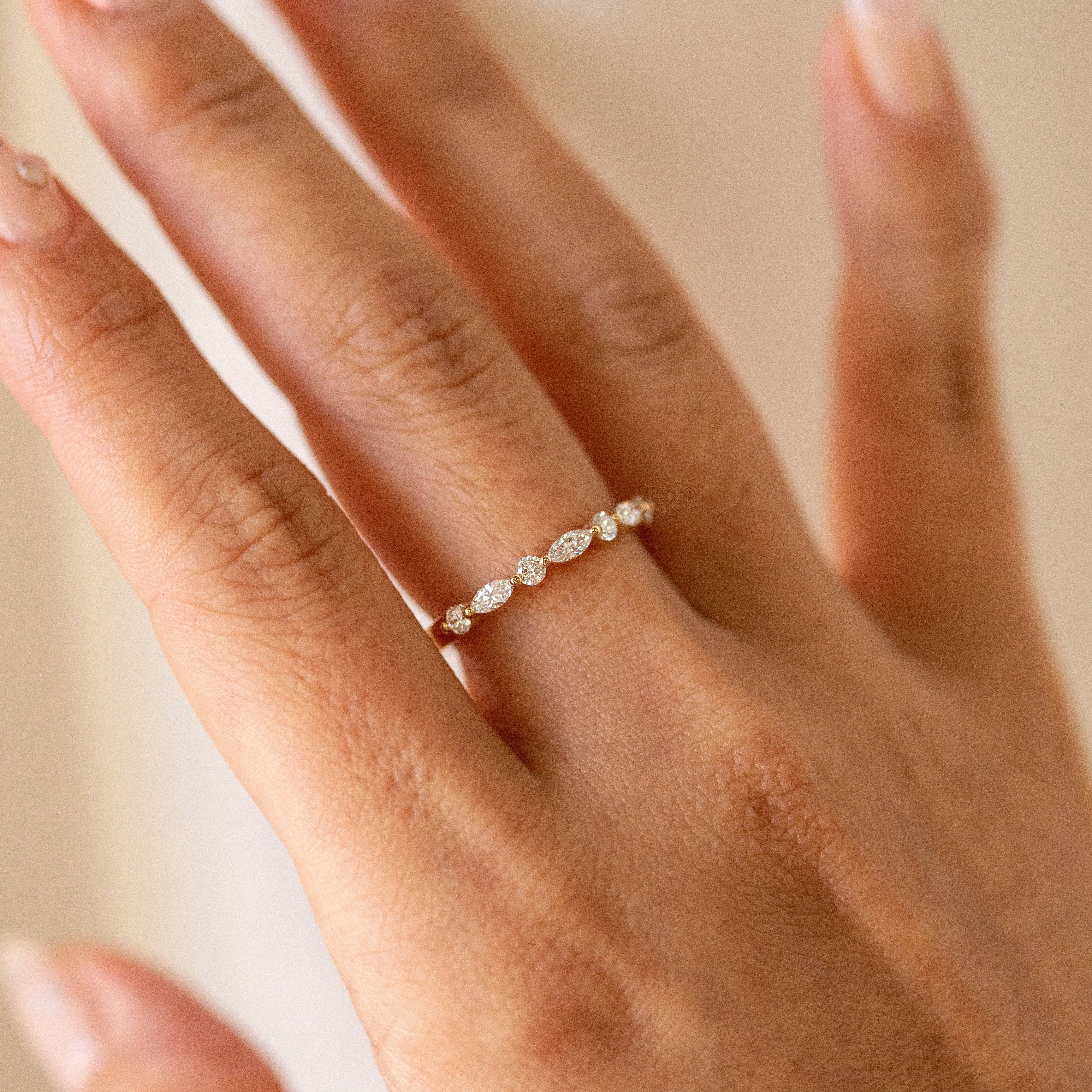A delicate gold band ring with a row of small clear marquise-cut diamond worn on a woman's ring finger. The hand has manicured nails with a light pink polish, set against a soft, warm beige background.