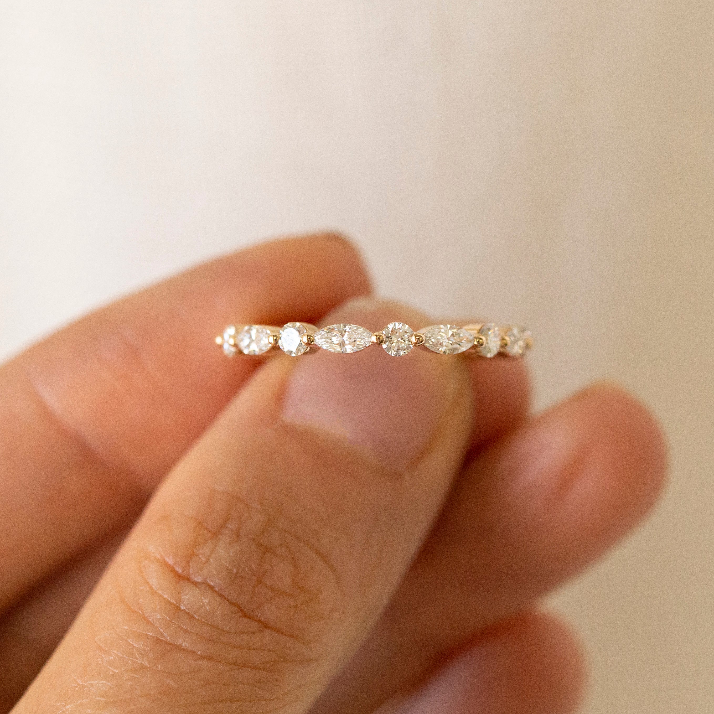 A delicate gold eternity ring with small round-cut clear stones set across the band, held between fingers against a soft beige background.