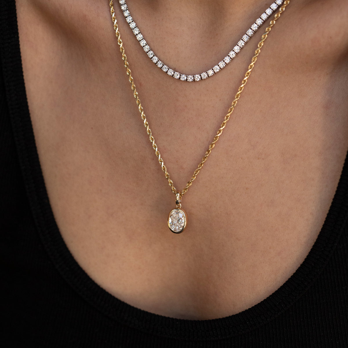 A close-up of a model's neck and chest wearing two layered necklaces: a gold chain with a teardrop-shaped clear gemstone pendant and a silver-toned tennis necklace.