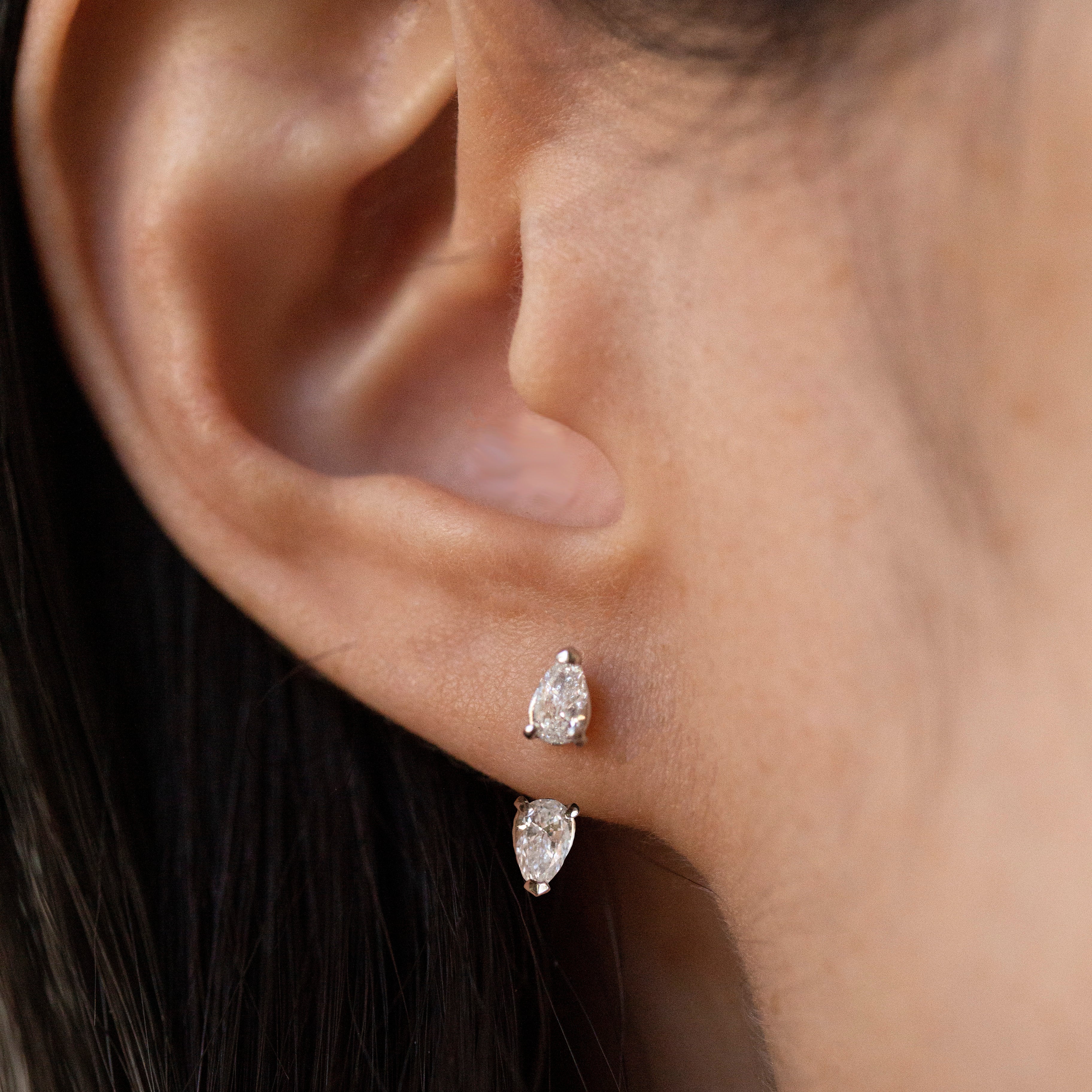 Close-up of a person's ear wearing two small pear-cut stud earrings in a silver-toned setting.