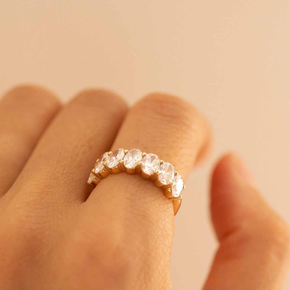 A close-up of a hand wearing a gold eternity ring set with clear round-cut gemstones, set against a solid warm beige background.