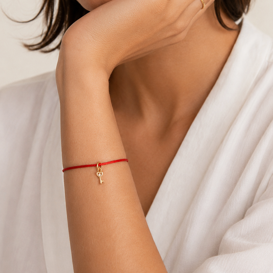 A close-up of a woman wearing a delicate red cord bracelet with a small gold key charm on her wrist, paired with a white robe.
