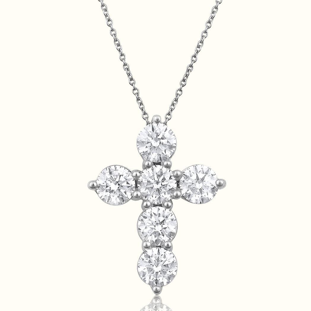 A silver cross pendant necklace set with round-cut clear gemstones, suspended from a delicate silver chain against a light cream background with a soft reflection below.
