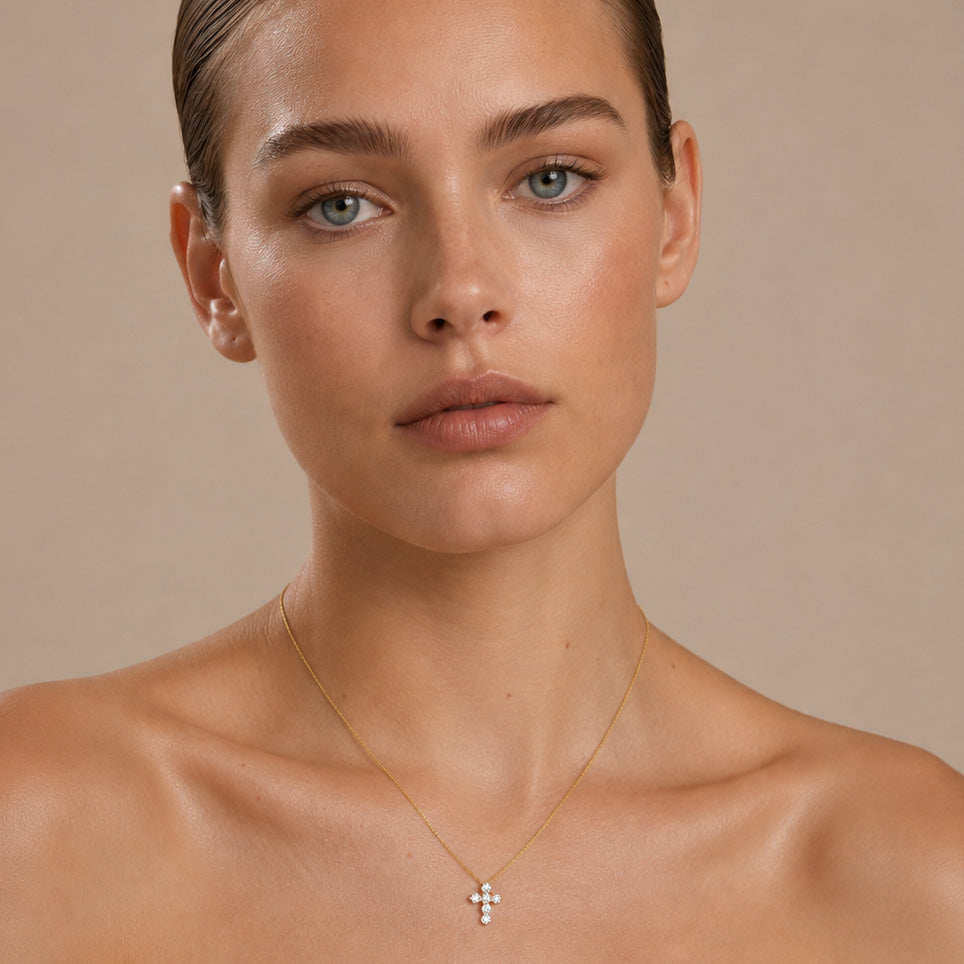 A close-up of a model wearing a delicate gold cross pendant necklace with small clear stones, set against a solid beige background.