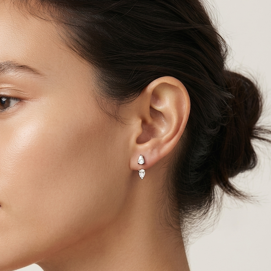 A close-up side profile of a woman wearing a pair of small PEAR DIAMOND earrings with clear stones, set against a clean, light beige background.