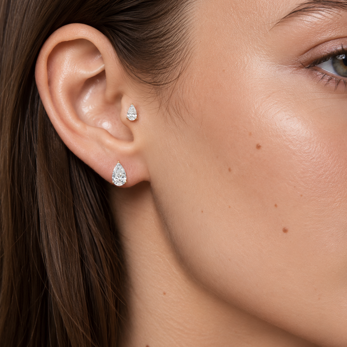 Close-up of a woman's ear wearing two small pear-shaped stud earrings with clear sparkling stones.