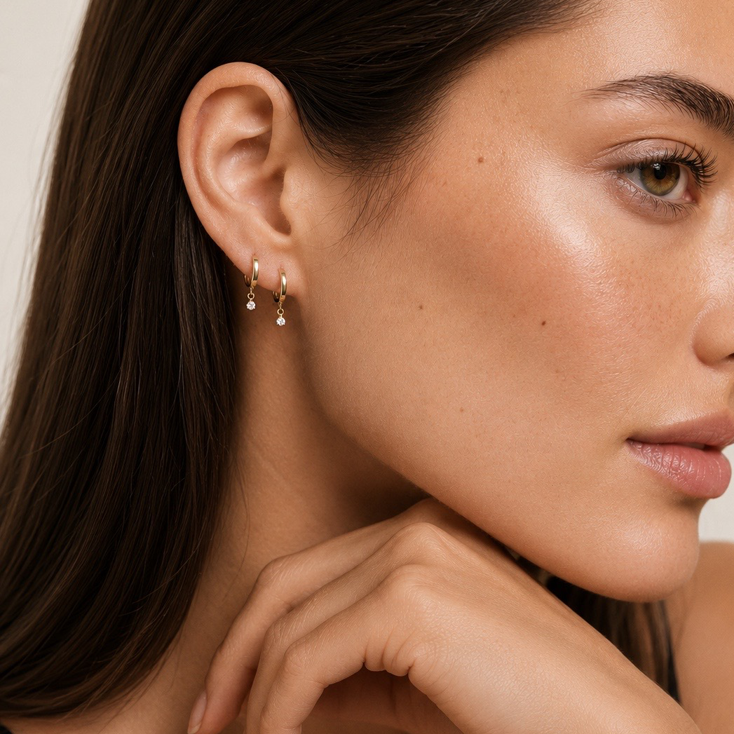 A close-up of a woman wearing a delicate gold huggie hoop earring with small dangling teardrop crystals, shown against a soft beige background.