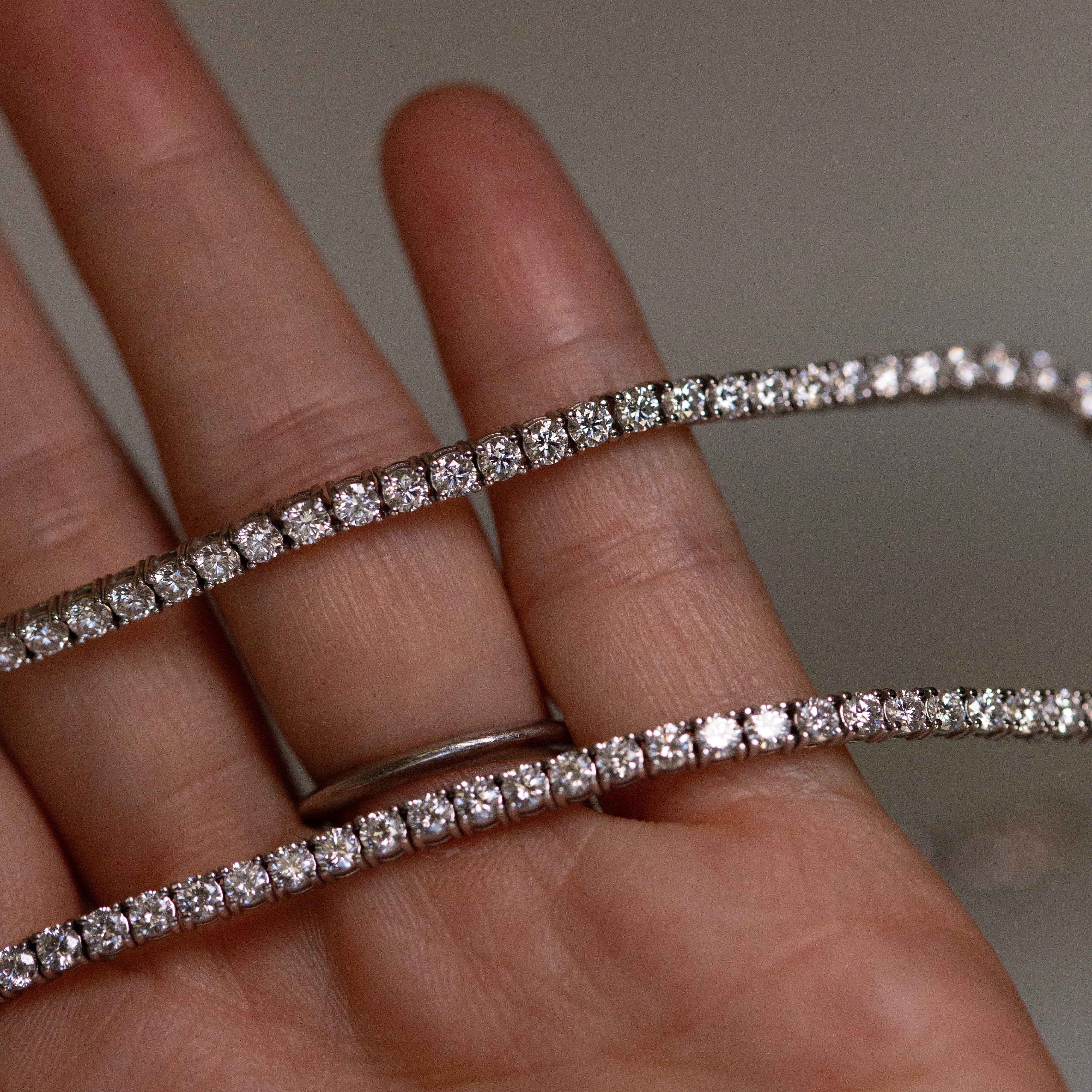 A close-up of a hand holding a delicate white gold tennis bracelet with brilliant diamonds, with a matching silver band visible on the ring finger.