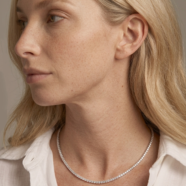 A close-up side profile of a blonde woman wearing a delicate silver tennis necklace and a white collared shirt against a soft beige background.