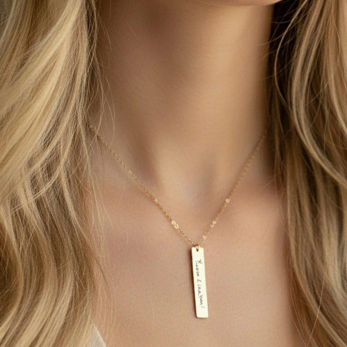 Gold necklace with a vertical bar pendant on a person with blonde hair.