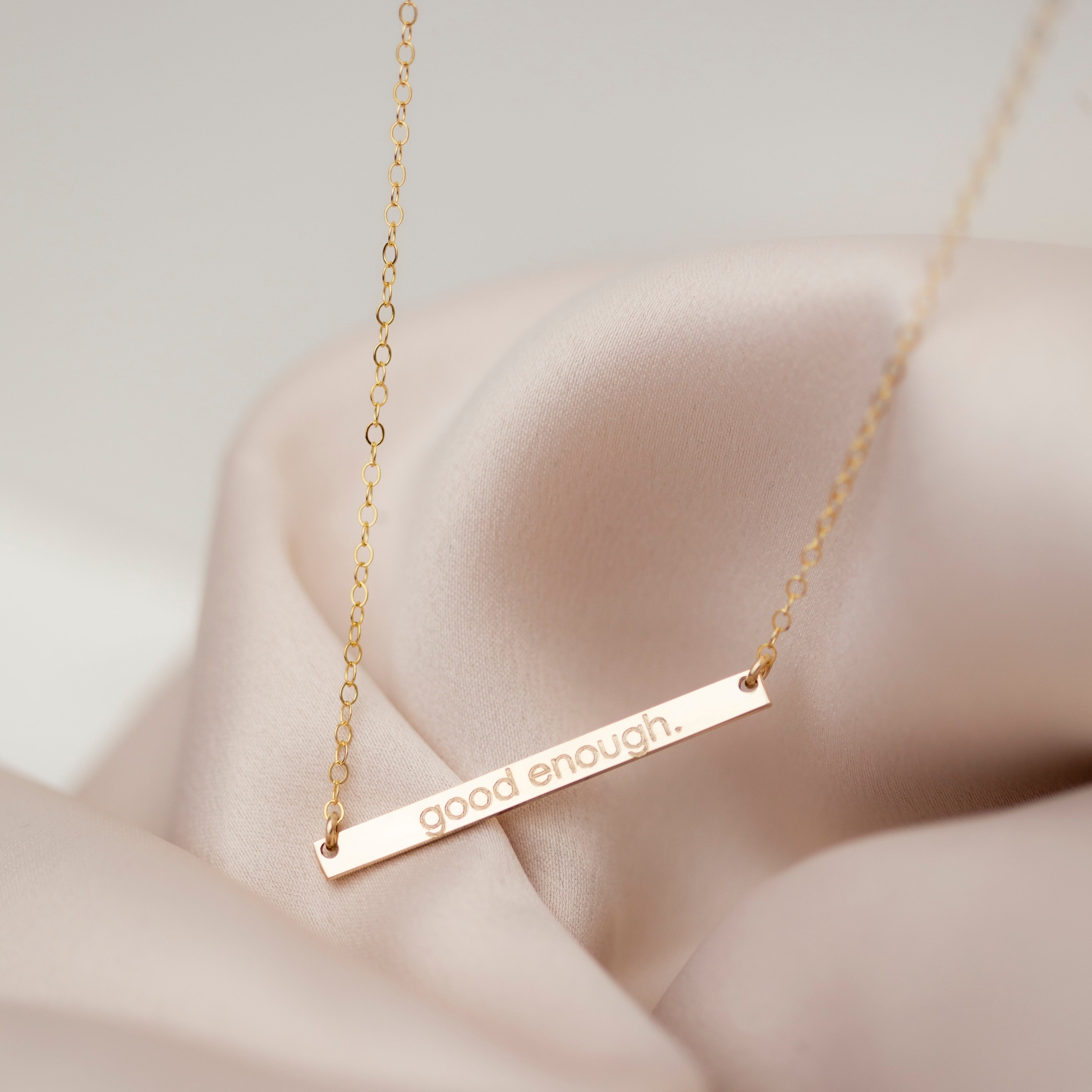 A gold-filled skinny name bar plate necklace laid out on a silky surface, with the bar featuring engraved personalized text.