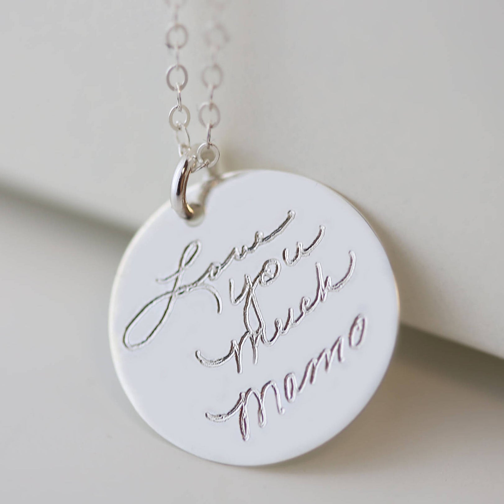 A silver necklace with a round pendant that says 'Love you much Mama' in cursive handwriting.