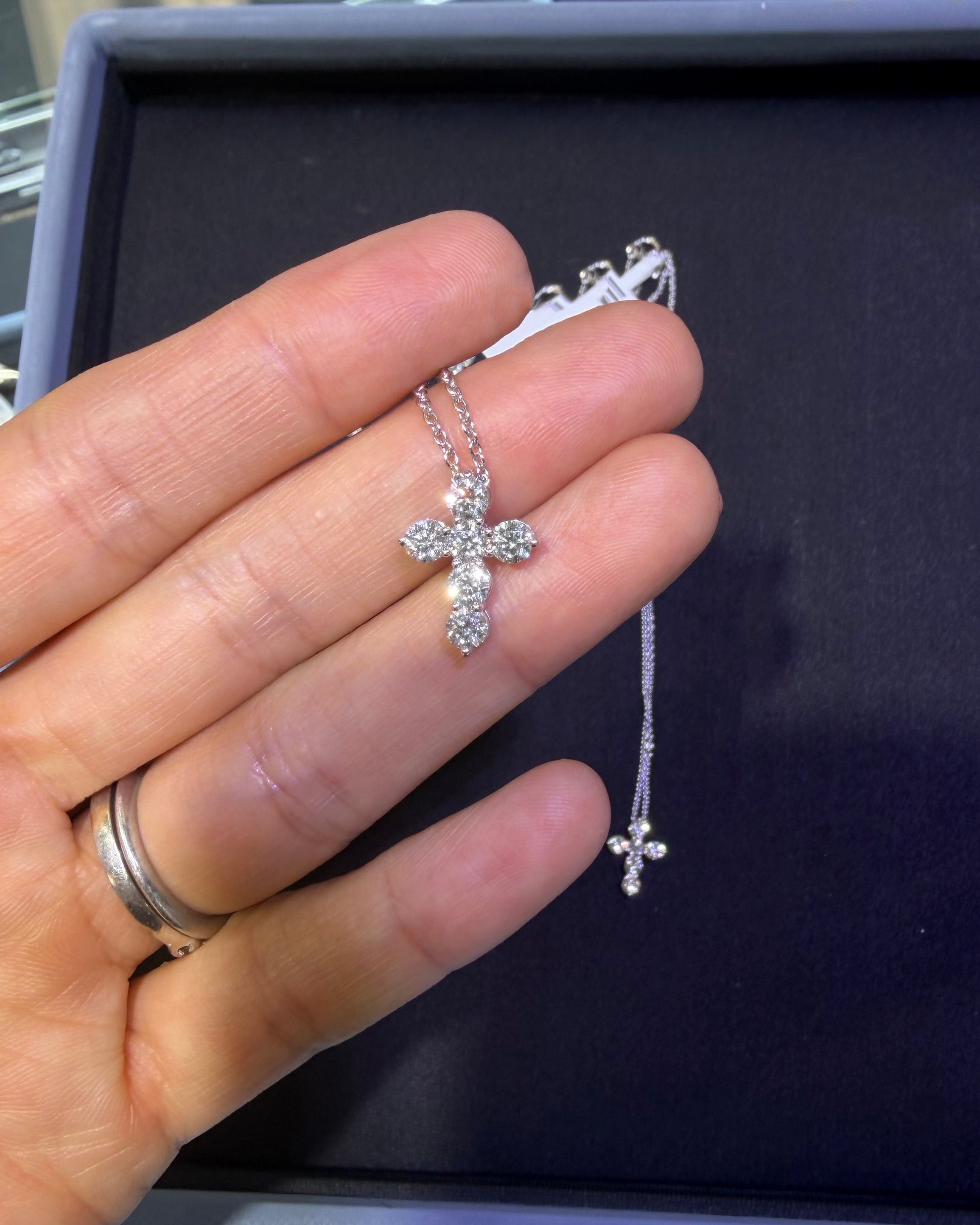 A silver cross pendant necklace with clear stones, held in a person's hand over a dark jewelry box.