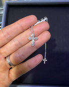 A silver cross pendant necklace with clear stones, held in a person's hand over a dark jewelry box.