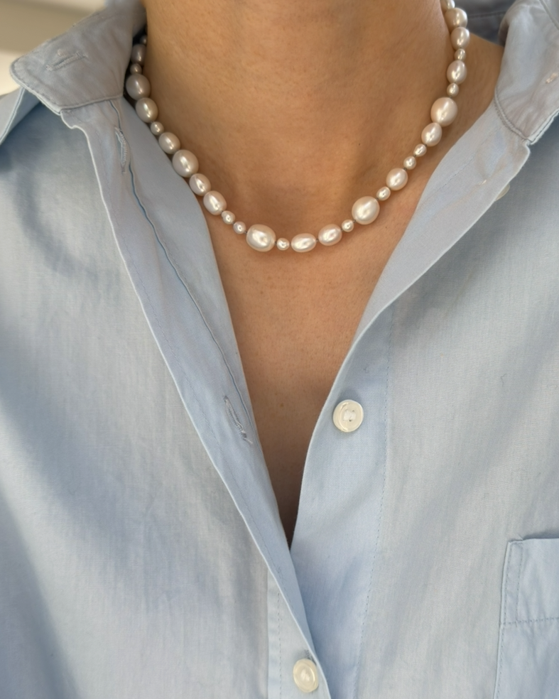 A close-up of a person wearing a double-strand pearl necklace over a light blue button-down shirt.