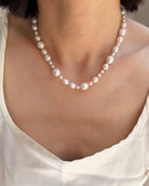 A close-up of a model wearing a pearl necklace with varying sizes of white pearls, paired with a white top.