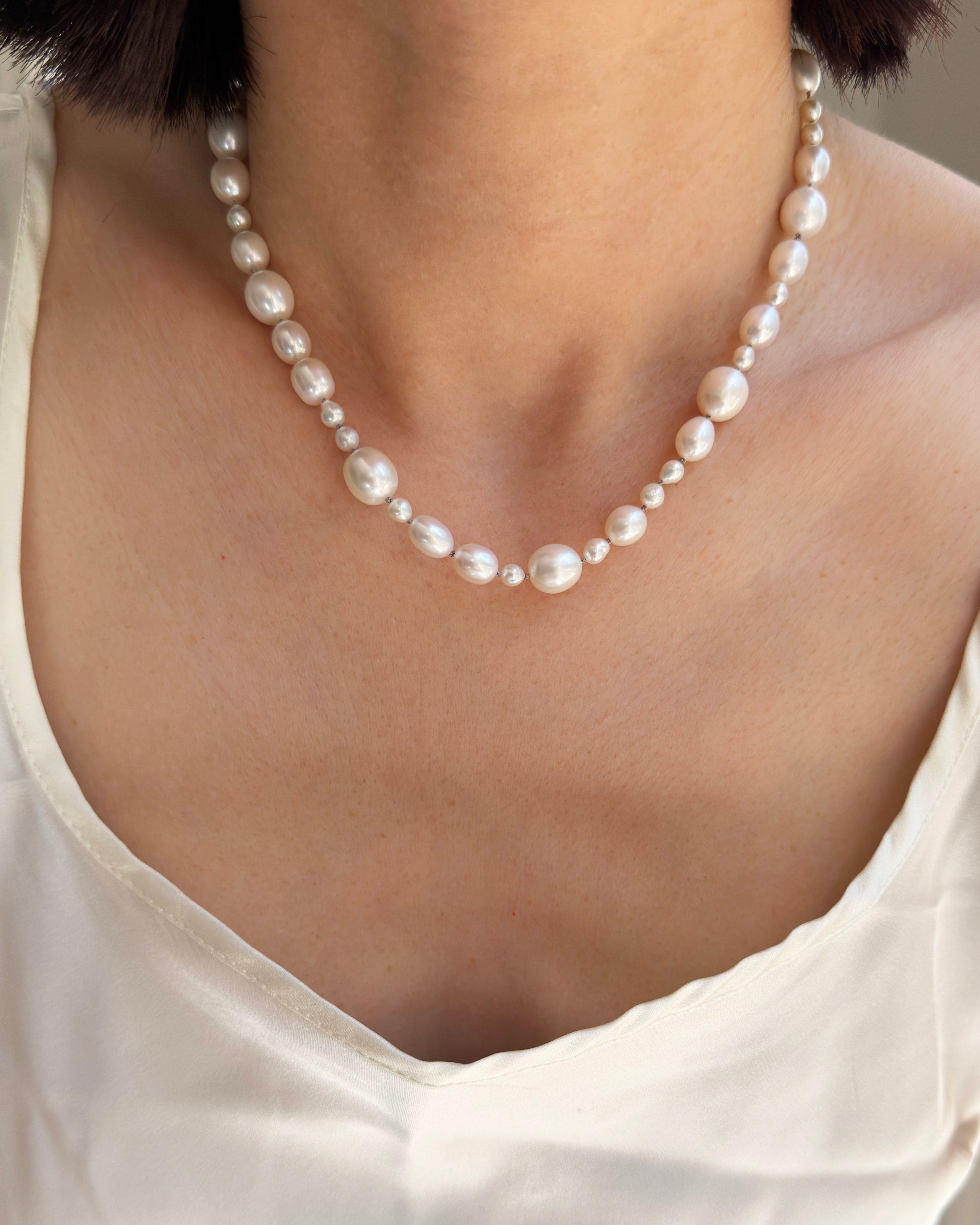 A close-up of a model wearing a pearl necklace with varying sizes of white pearls, paired with a white top.