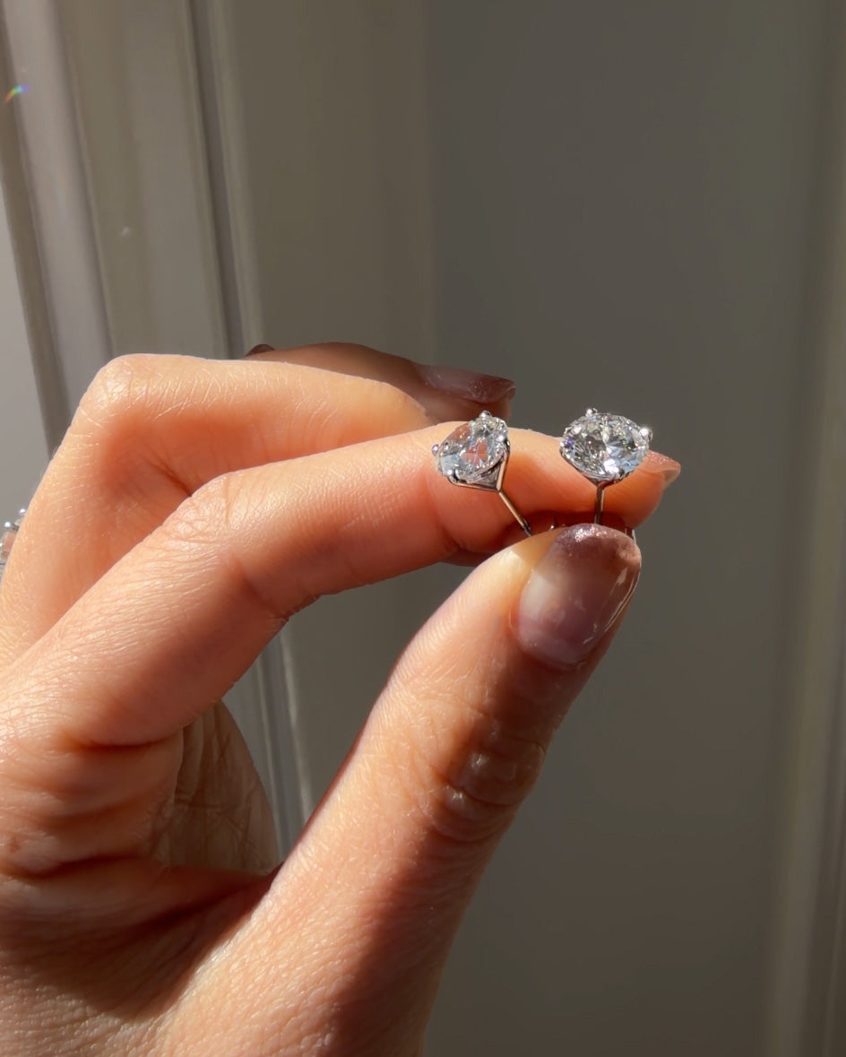 Hand holding two diamond stud earrings against a neutral background
