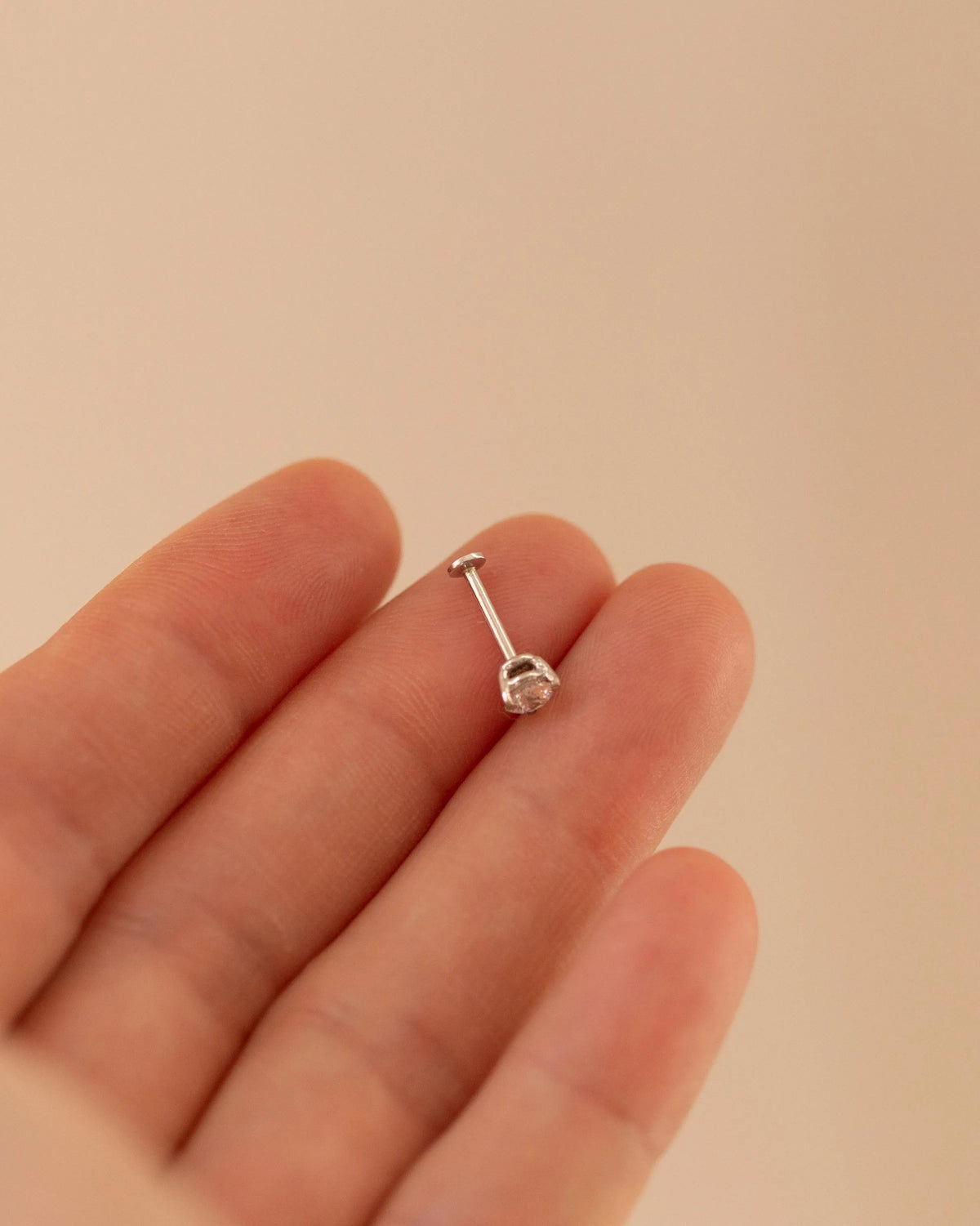 A silver-toned labret piercing stud with a clear gemstone, shown resting on a person's fingers against a soft beige background.