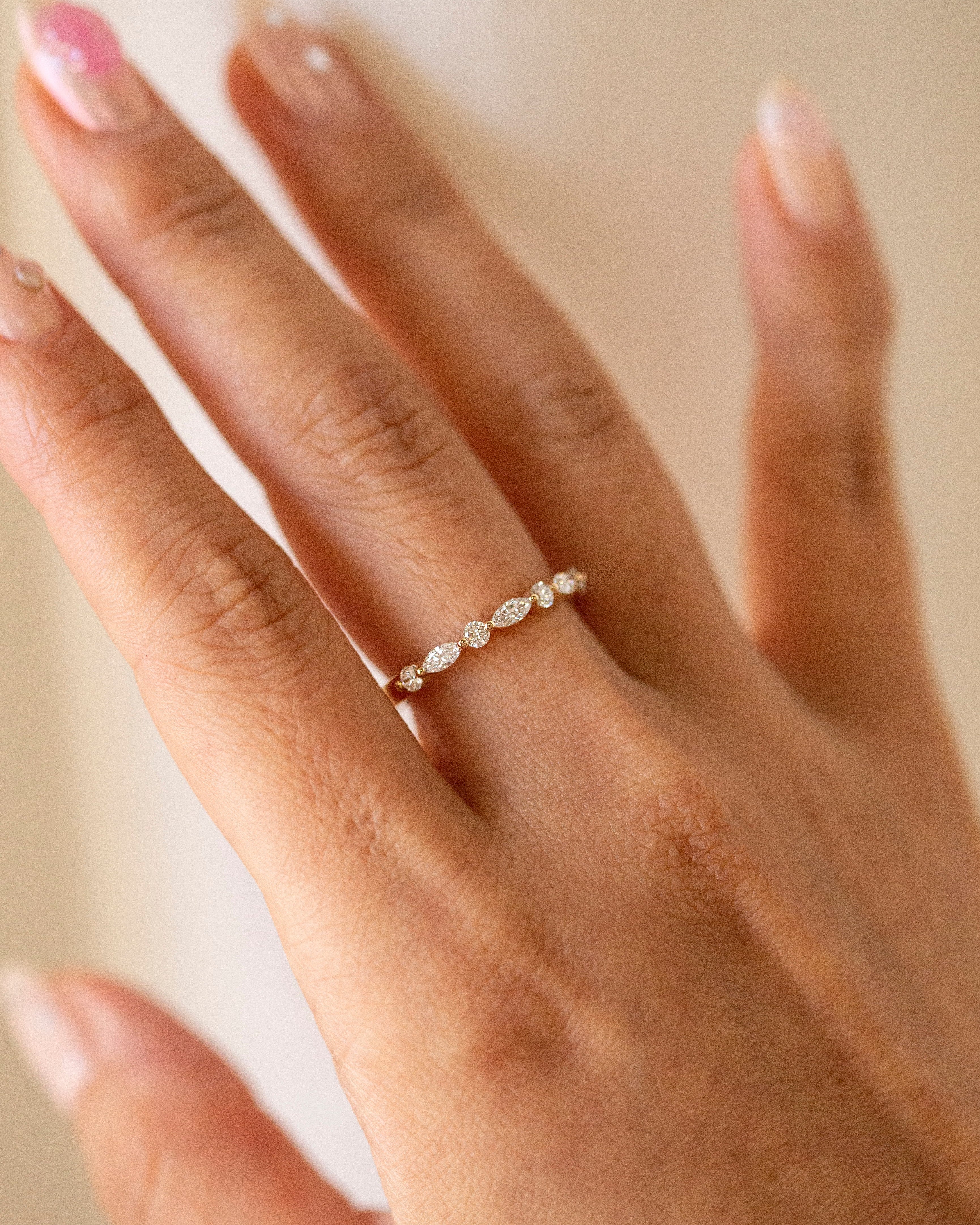 A delicate gold band ring with a row of small clear marquise-cut diamond worn on a woman's ring finger. The hand has manicured nails with a light pink polish, set against a soft, warm beige background.