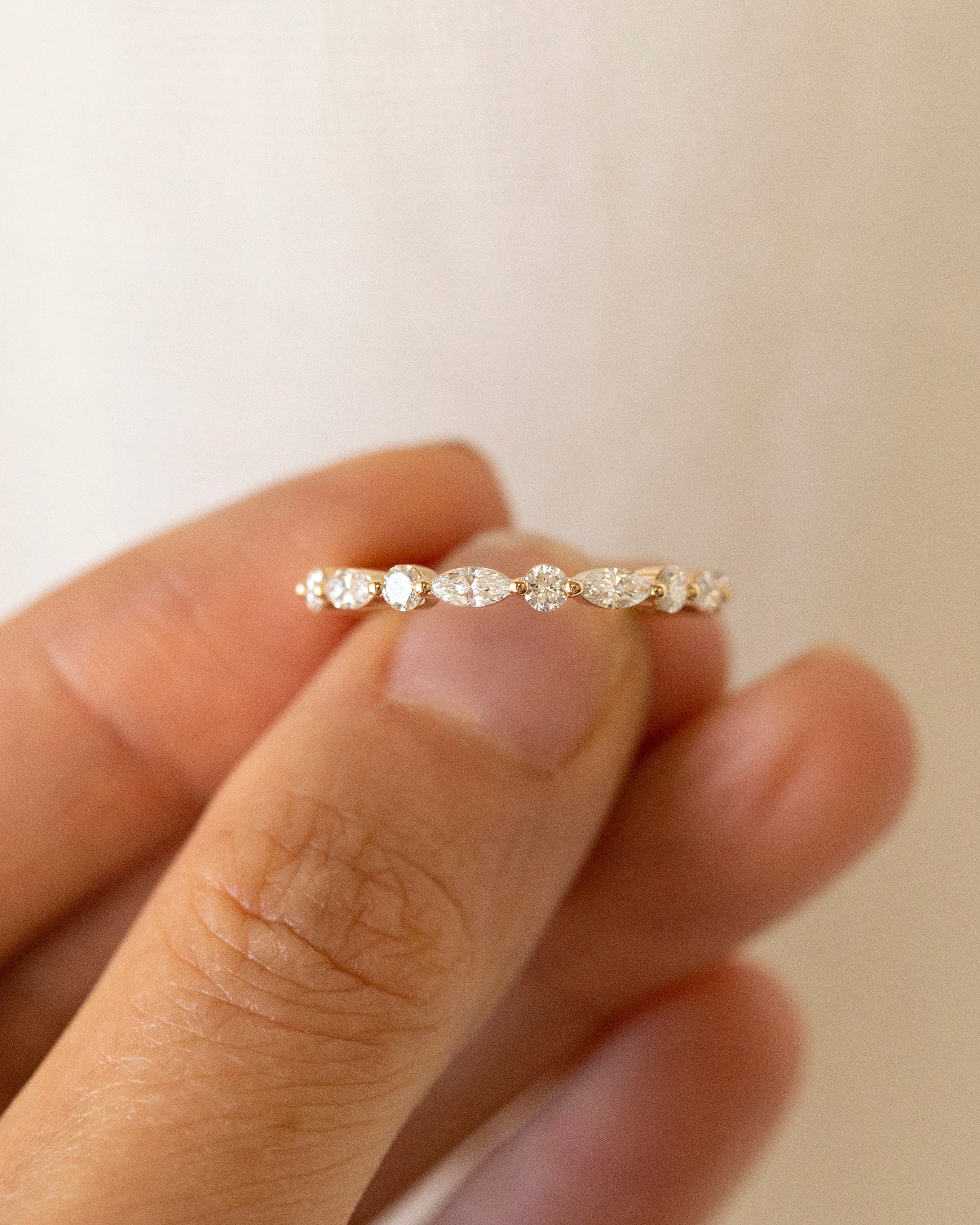 A delicate gold eternity ring with small round-cut clear stones set across the band, held between fingers against a soft beige background.