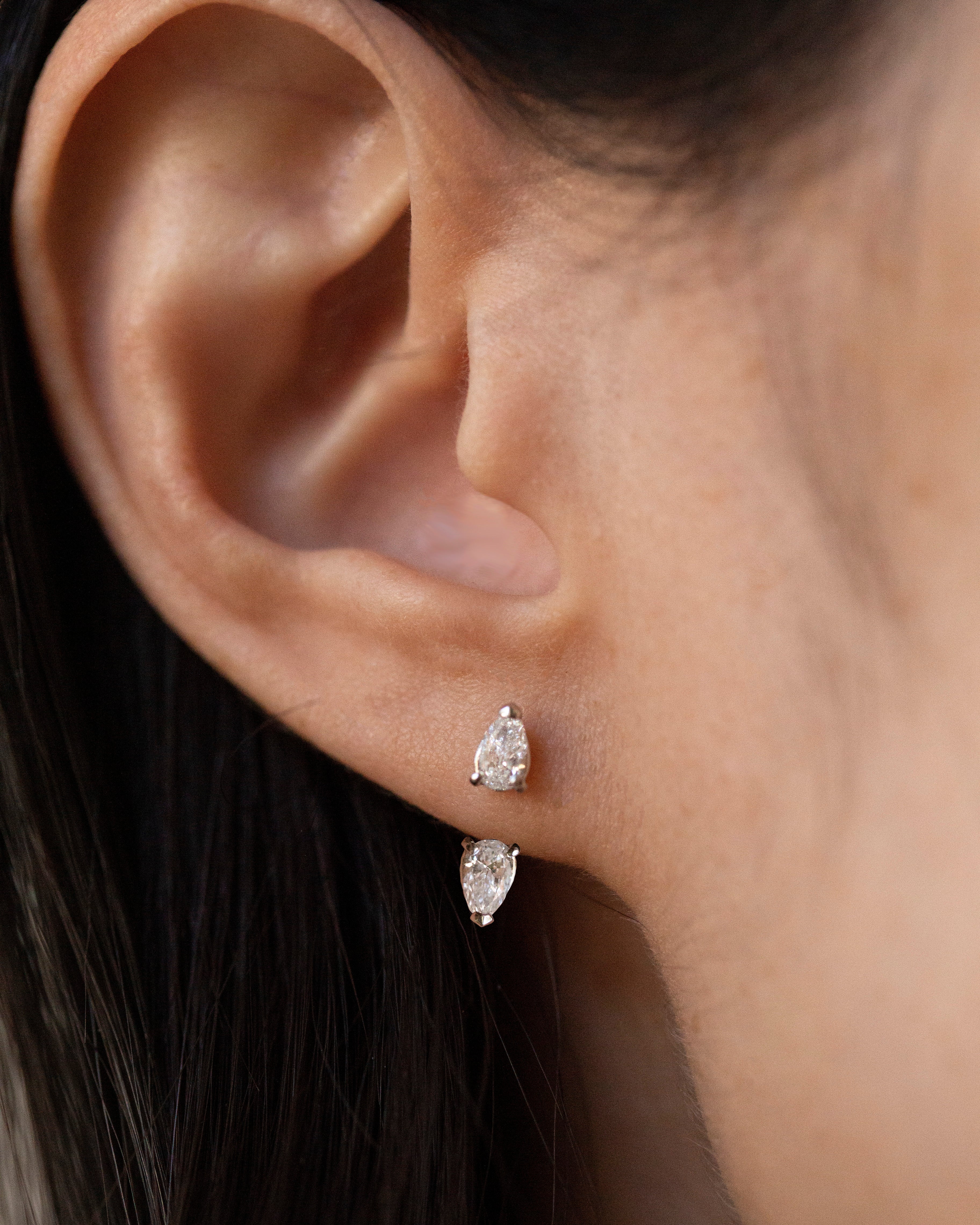 Close-up of a person's ear wearing two small pear-cut stud earrings in a silver-toned setting.