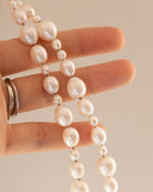 A close-up of a hand holding a strand of irregular white freshwater pearls with silver spacer beads, against a solid beige background.
