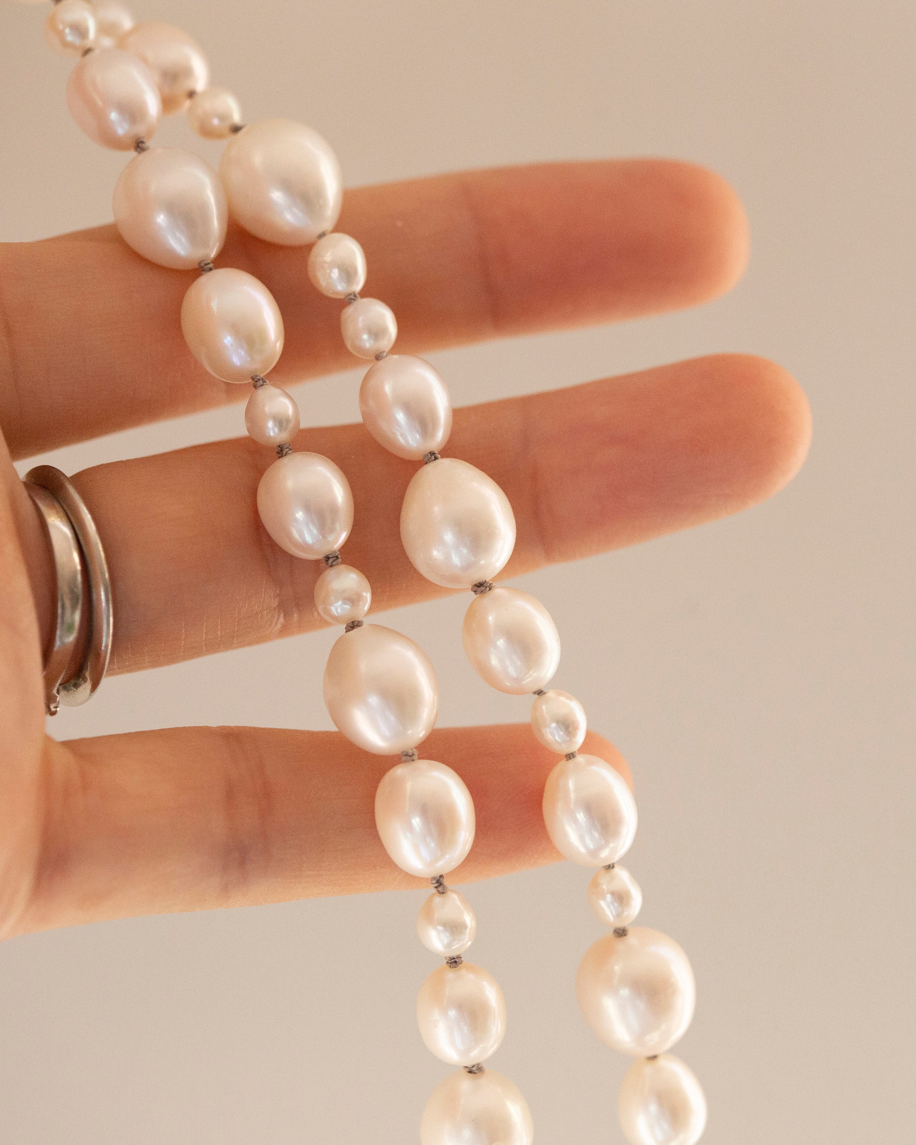 A close-up of a hand holding a strand of irregular white freshwater pearls with silver spacer beads, against a solid beige background.