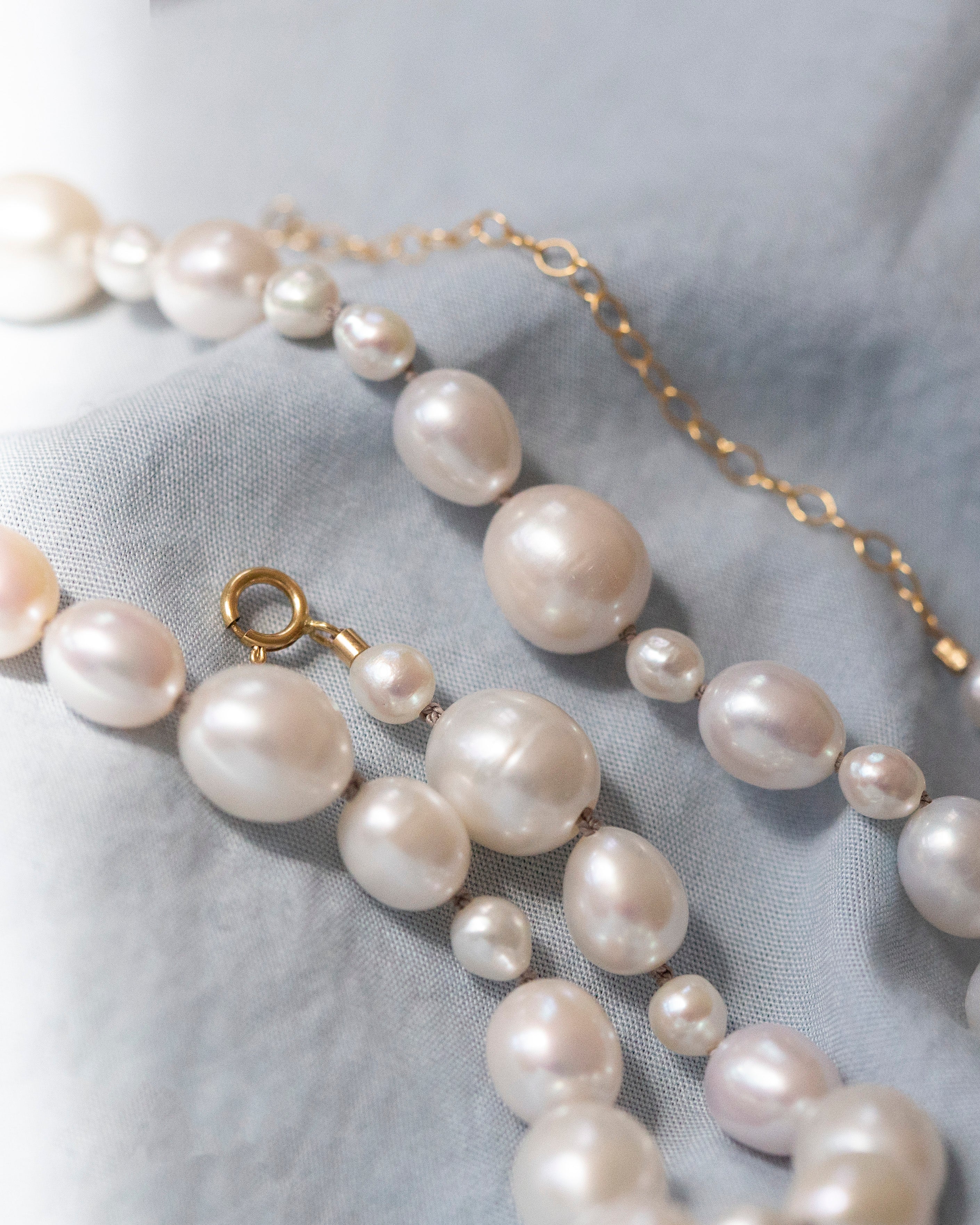 A close-up of a pearl necklace with irregular baroque-style pearls in soft white and pale pink tones, accented by a gold chain extender and clasp, laid on a textured light gray fabric background.