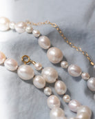 A close-up of a pearl necklace with irregular baroque-style pearls in soft white and pale pink tones, accented by a gold chain extender and clasp, laid on a textured light gray fabric background.