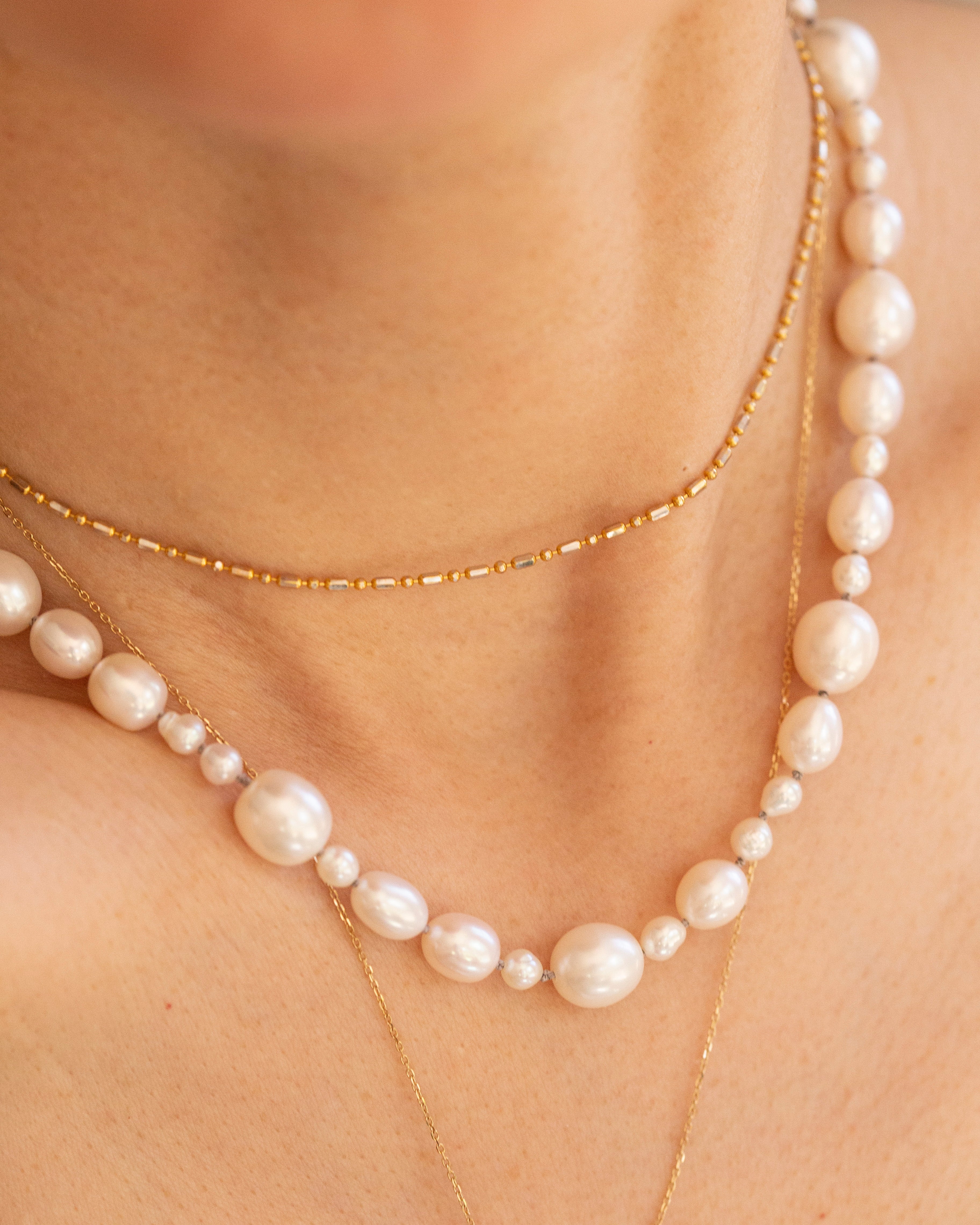 A close-up of a person's neck and upper chest wearing layered necklaces: a strand of large white pearls with small gold beads, a delicate gold chain, and a longer gold chain layered underneath.