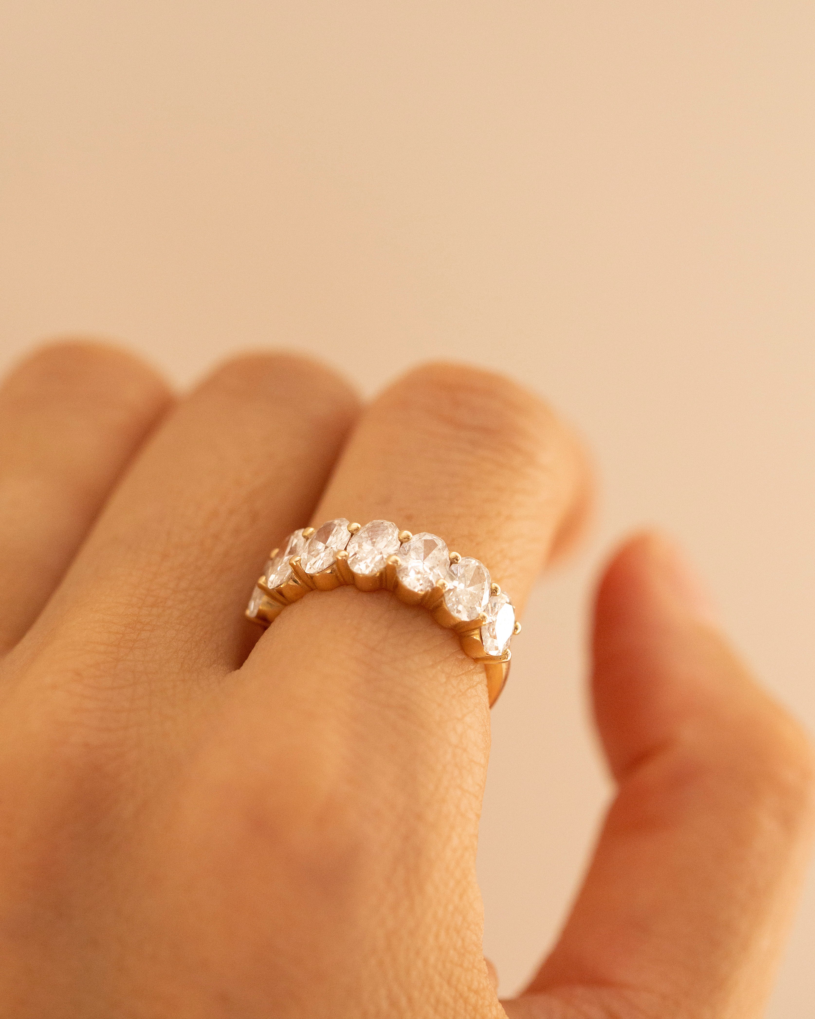 A close-up of a hand wearing a gold eternity ring set with clear round-cut gemstones, set against a solid warm beige background.