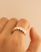 A close-up of a hand wearing a gold eternity ring set with clear round-cut gemstones, set against a solid warm beige background.