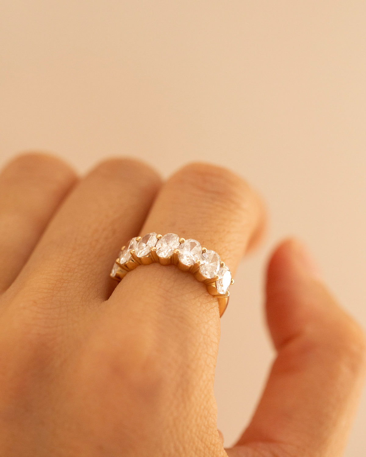 A close-up of a hand wearing a gold eternity ring set with clear round-cut gemstones, set against a solid warm beige background.