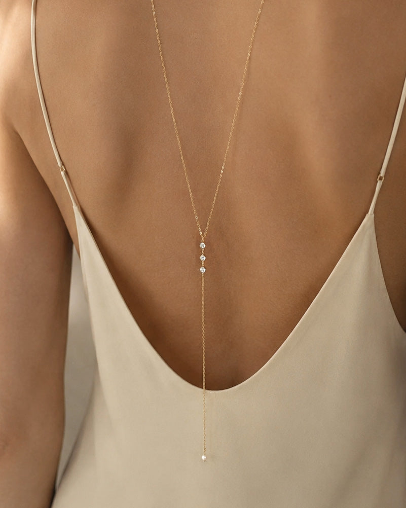 A delicate gold Y-necklace with three small white pearl-like beads at the center, shown on a model's back wearing a beige open-back camisole.