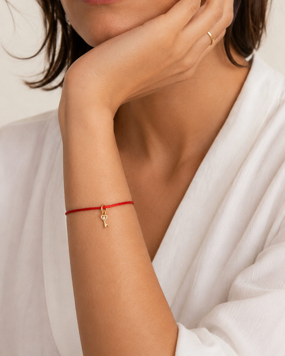 A close-up of a woman wearing a delicate red cord bracelet with a small gold key charm on her wrist, paired with a white robe.