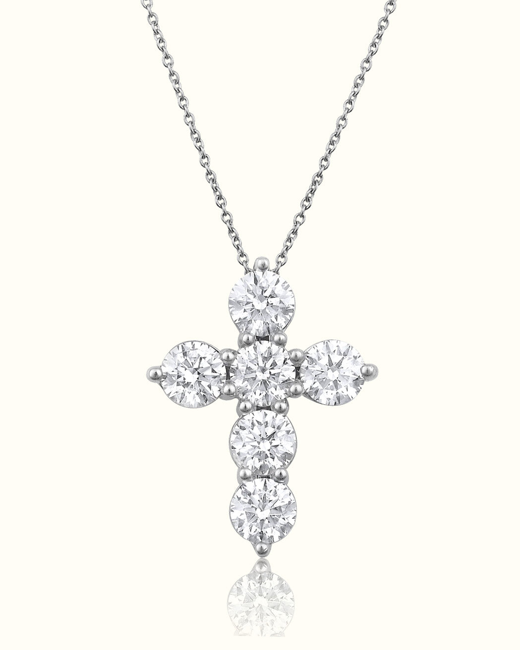 A silver cross pendant necklace set with round-cut clear gemstones, suspended from a delicate silver chain against a light cream background with a soft reflection below.