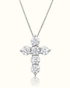 A silver cross pendant necklace set with round-cut clear gemstones, suspended from a delicate silver chain against a light cream background with a soft reflection below.