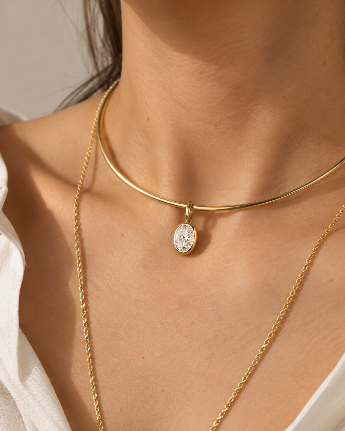 A close-up of a model's neck and chest wearing a gold bangle necklace with an oval clear gemstone pendant, layered over a longer gold chain necklace.