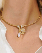 A close-up of a model's neck wearing a gold bangle necklace with decorative charms, including a teardrop crystal pendant and a sunburst disc charm.