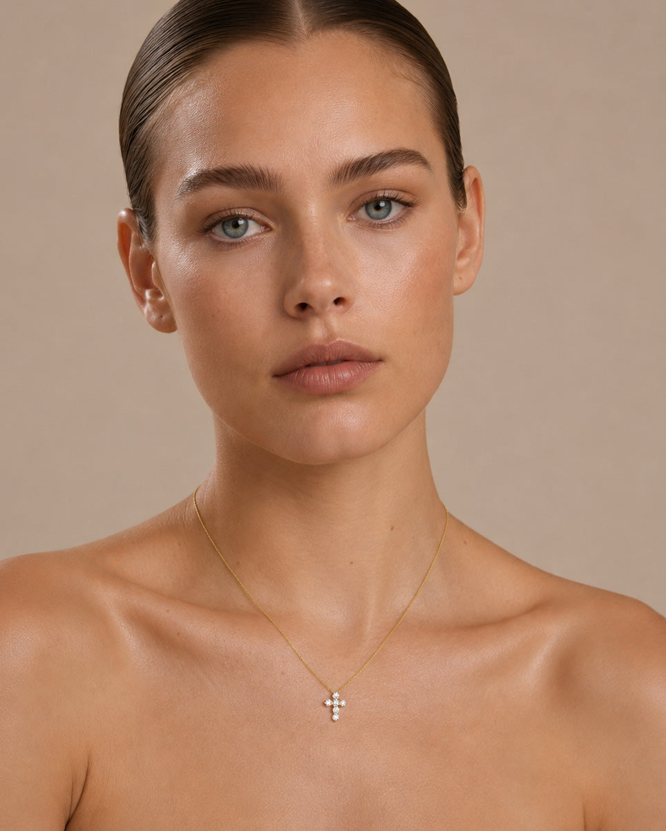 A close-up of a model wearing a delicate gold cross pendant necklace with small clear stones, set against a solid beige background.