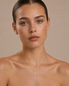 A close-up of a model wearing a delicate gold cross pendant necklace with small clear stones, set against a solid beige background.