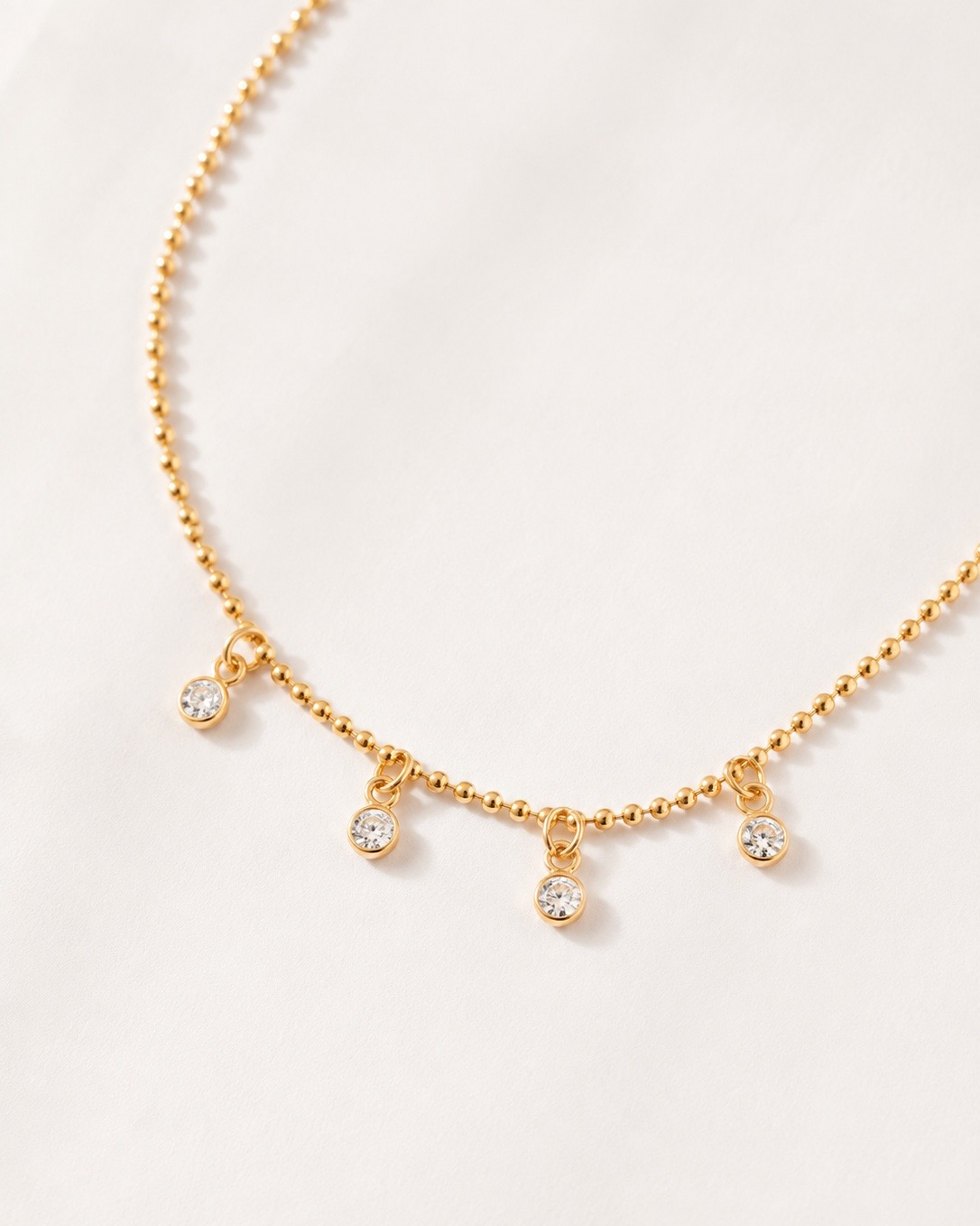 A delicate gold beaded necklace with four small, clear crystal dangle charms, displayed on a soft, light cream fabric background.