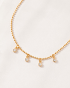 A delicate gold beaded necklace with four small, clear crystal dangle charms, displayed on a soft, light cream fabric background.