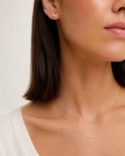Close-up of a woman wearing delicate layered gold necklaces and a small stud earring against a soft light background.